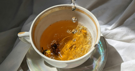 Revive & Thrive The Benefits of Cleansing. Image features herbal tea being poured into a small white mug. Herbs can be seen floating throughout the tea and an iridescent reflection shines off the saucer for the mug.