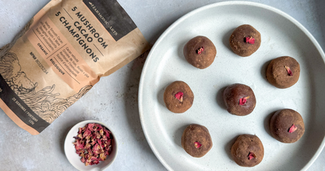 Image features a bag of Harmonic Arts 5 Mushroom Cacao next to a plate of Rose Cacao Truffles. There is a small bowl of rose petals between the bag and the plate.